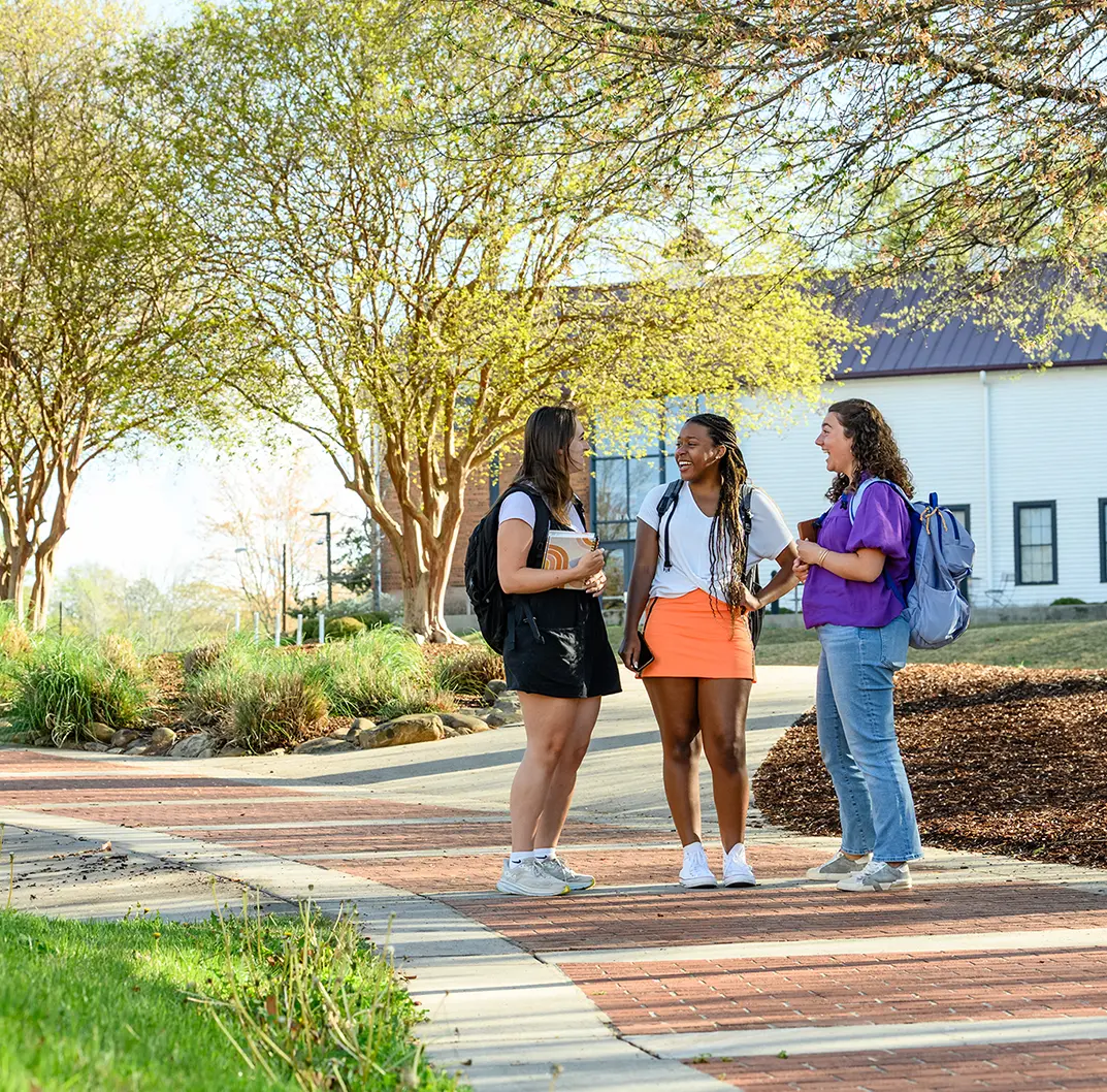 Three female students with backpacks stand on a walkway in the middle of campus in front of a painted white and brick building and green leafy trees.