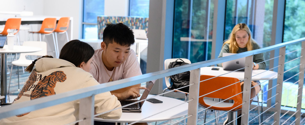 a male and female student look at a laptop together in the humanities hall while other students study in the background