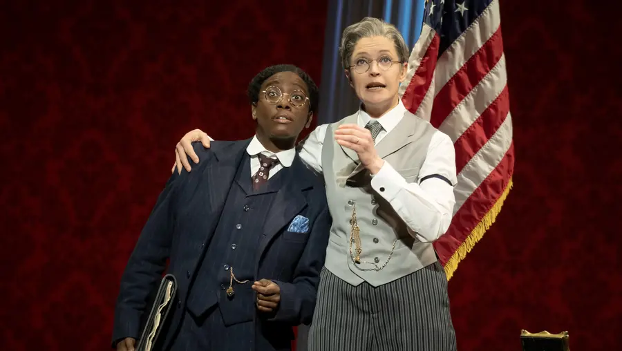 In 1920s-era costumes, a white woman dressed as a man with a moustache speaks with her arm around a Black woman dressed as a man, who looks reluctant to the companion's message. The pair stand in front of an American flag on a stand.