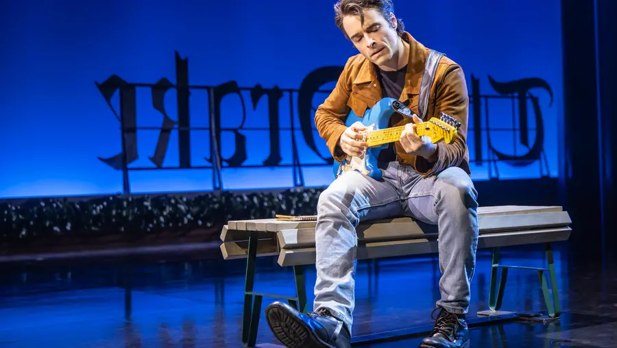 Man wearing jeans and a brown leather jacket sings and plays a blue and white electric guitar on a stage set to look like a rooftop.