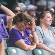 A person in a purple Rockies T-shirt sits in a stadium, one arm crossed and the other extended to scratch her head.