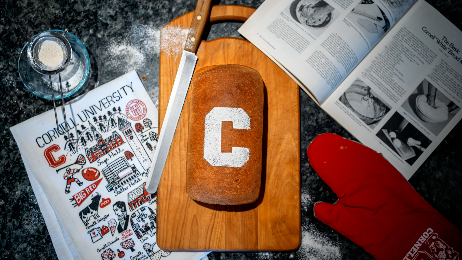 A counter with cooking implements and a fresh loaf of bread with a 'C' sifted on top.