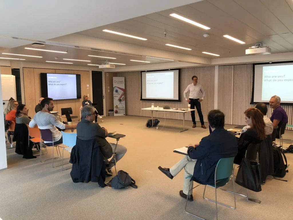 A man standing in a room presenting to a group of people sat on chairs in a U configuration