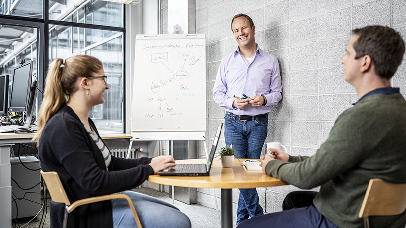 Ein Beschäftigter erklärt zwei Kolleg*innen einen Prozess, für die Darstellung nutzt er ein Flipchart.