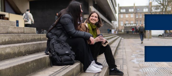 2 students sit on steps in front of main library - e in coloured blocks on right