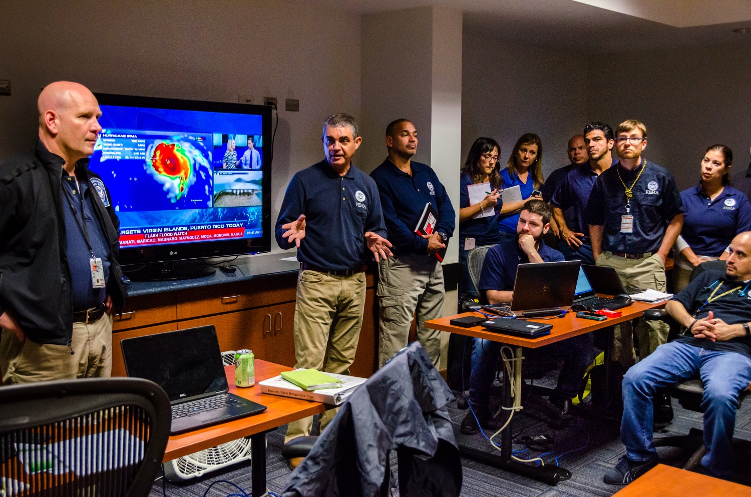  Room packed with FEMA employees watching a hurricane report on TV.