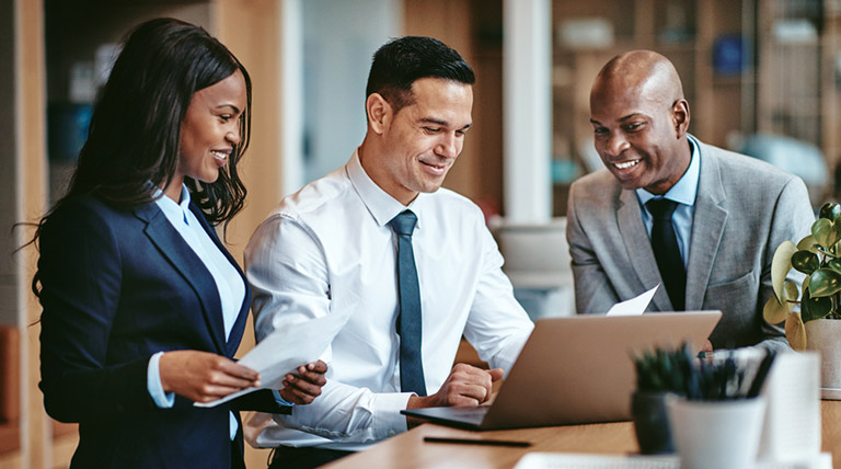 Three people in business attire smiling and looking at a laptop together at a desk