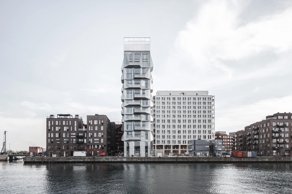 A photograph showing a dense group of modern buildings along a body of water. The central building is a tall, slim tower with irregular balconies protruding from its sides.