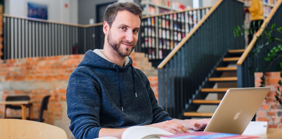 Student der Verwaltungsökonom in der Bibliothek am Fachbereich Verwaltungswissenschaften der Hochschule Harz blickt in die Kamera. Vor ihm liegen viele Bücher und ein Laptop