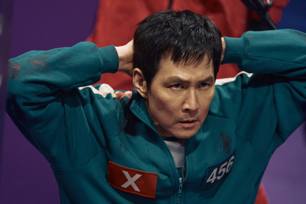 Squid Game Season 2 stars Lee Jung-jae as Seong Gi-hun, shown here with his hands behind his head, a guard standing behind him