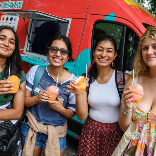 Four students smile while posing with cups of colorful Italian Ice; a red Italian Ice truck is behind them.