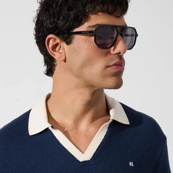 Man wearing sunglasses and a navy sweater with a white collar against a light background