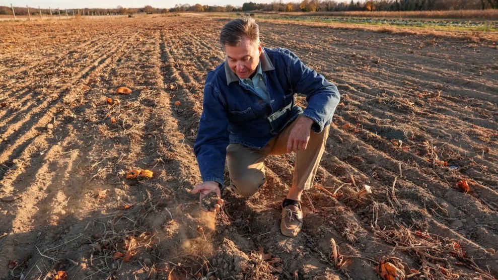 A white man wearing a blue jacket crouches in the soil.