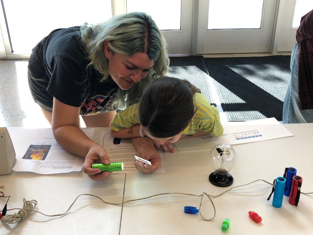 A parent and child lean in closely over a table to look at a flashlight shining at a small piece of paper.