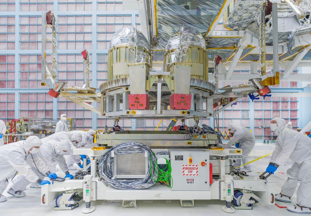 people in white, full-body "bunny suits" position hardware in a gleaming metallic clean room at NASA Goddard