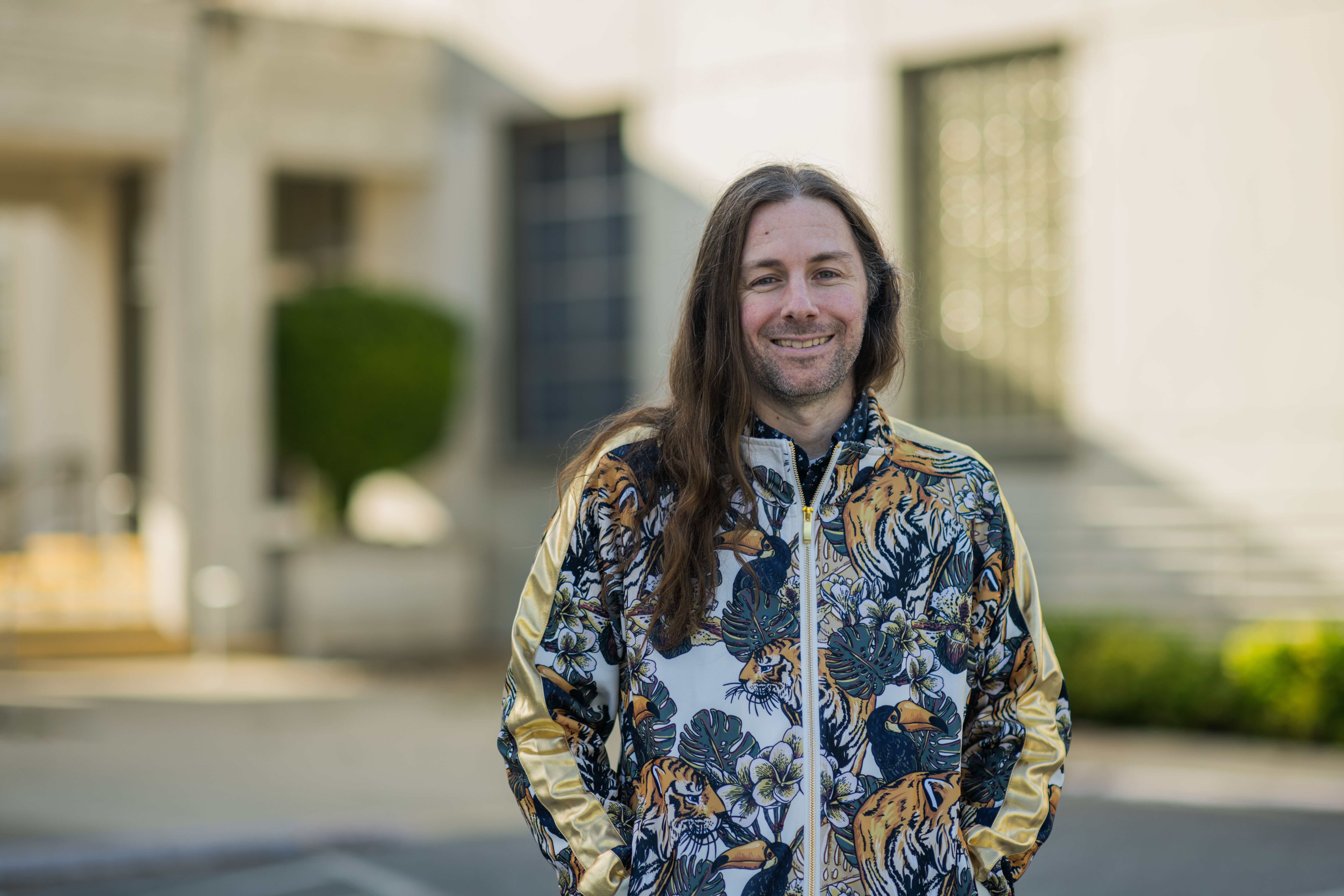 Garrett Sadler smiles softly outside in front of a marble building for his Faces of NASA portrait. His brown hair is long, past his shoulders, and he's wearing a zipped-up jacket with tropical plants, flowers, and tigers on it.