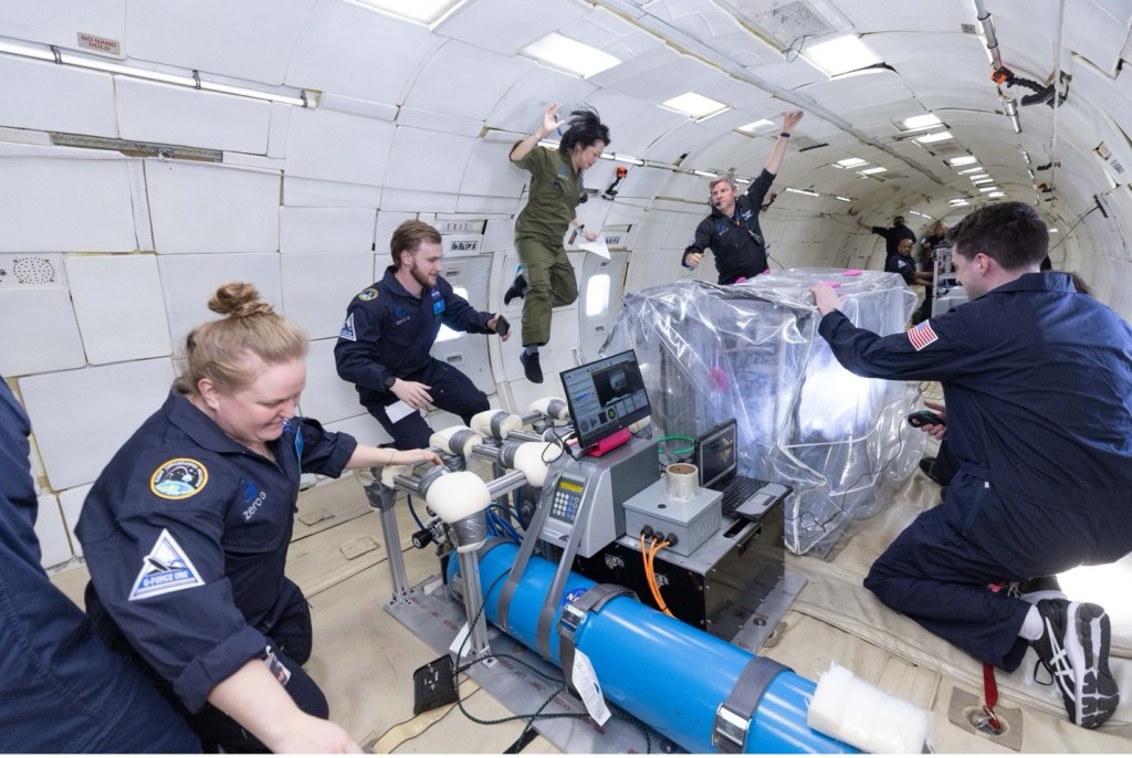 Five people in flight suits work on an experiment while floating in a simulated lunar gravity environment.