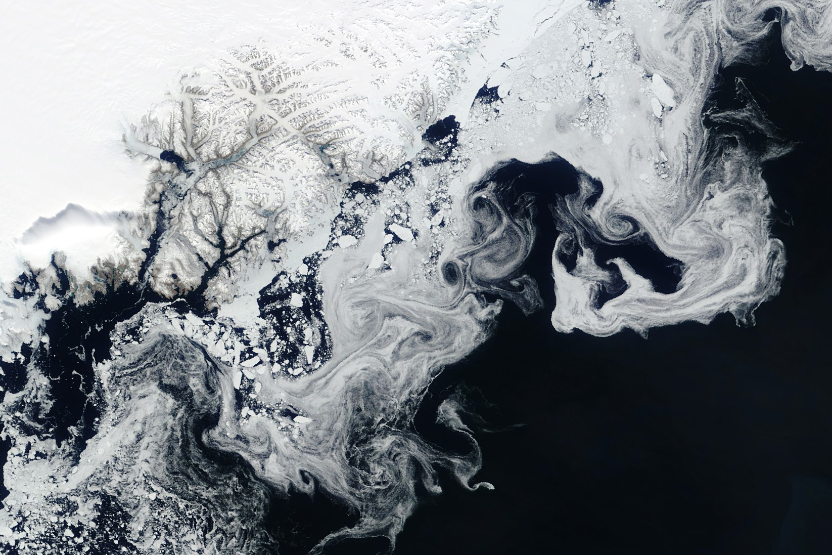 A satellite view of sea ice. The ice is white and solid at top left, while the edges swirl and swoop through the dark blue water.