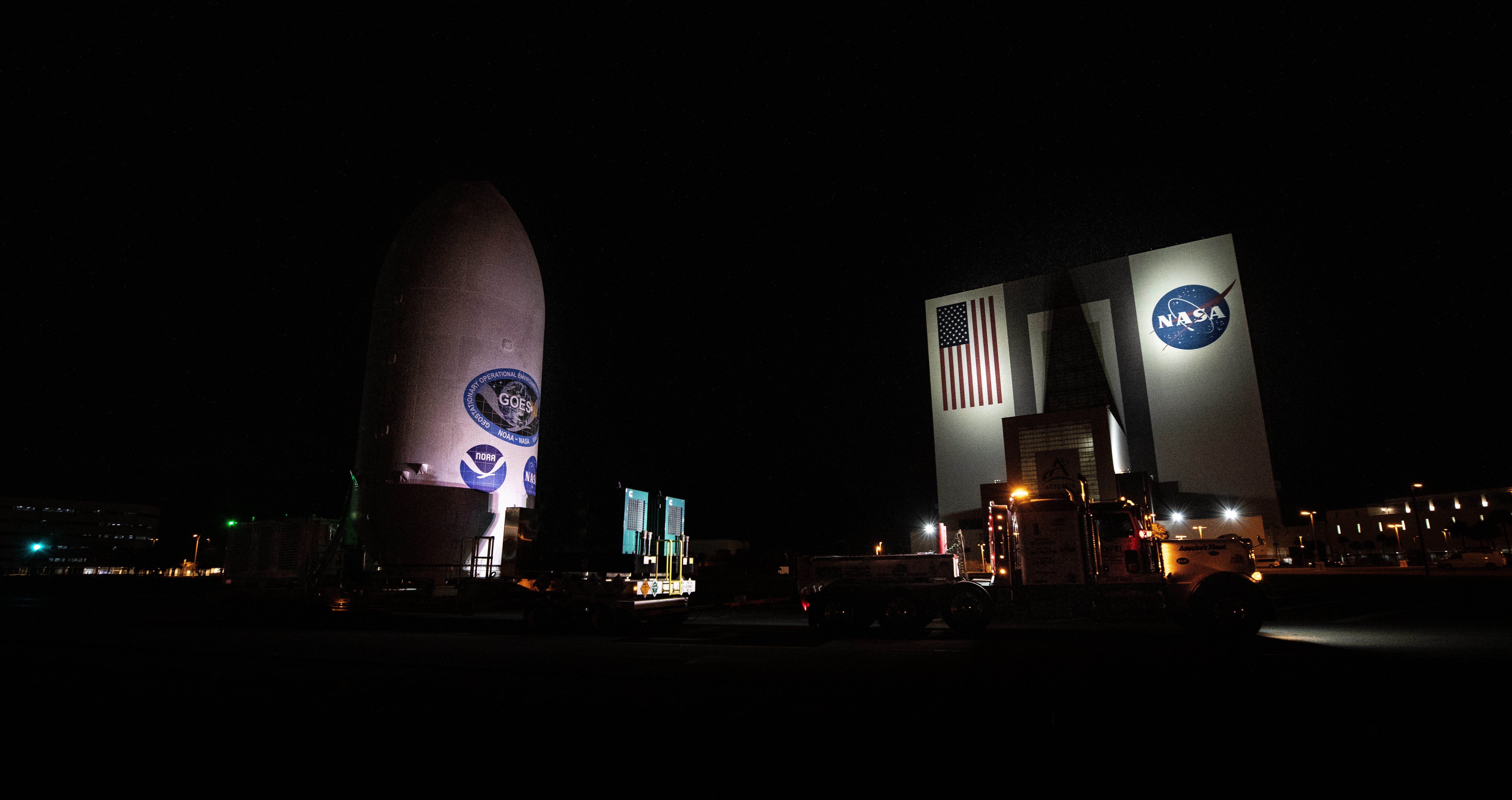 NOAA's GOES-U satellite (left) and NASA's Vehicle Assembly Building (right) are both lit up at night. GOES-U is a white cylindrical object and the VAB is a tall, square building.