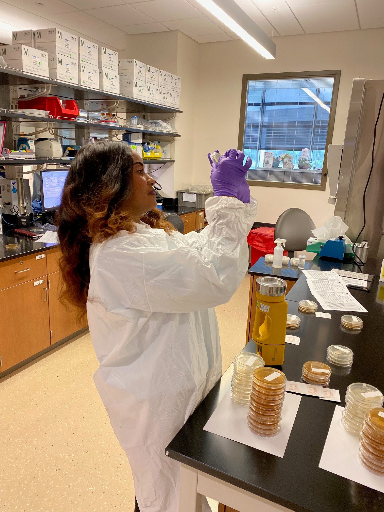 A woman in a white lab coat and purple gloves works in a laboratory. She is carefully holding a small vial or container up to eye level, examining its contents.