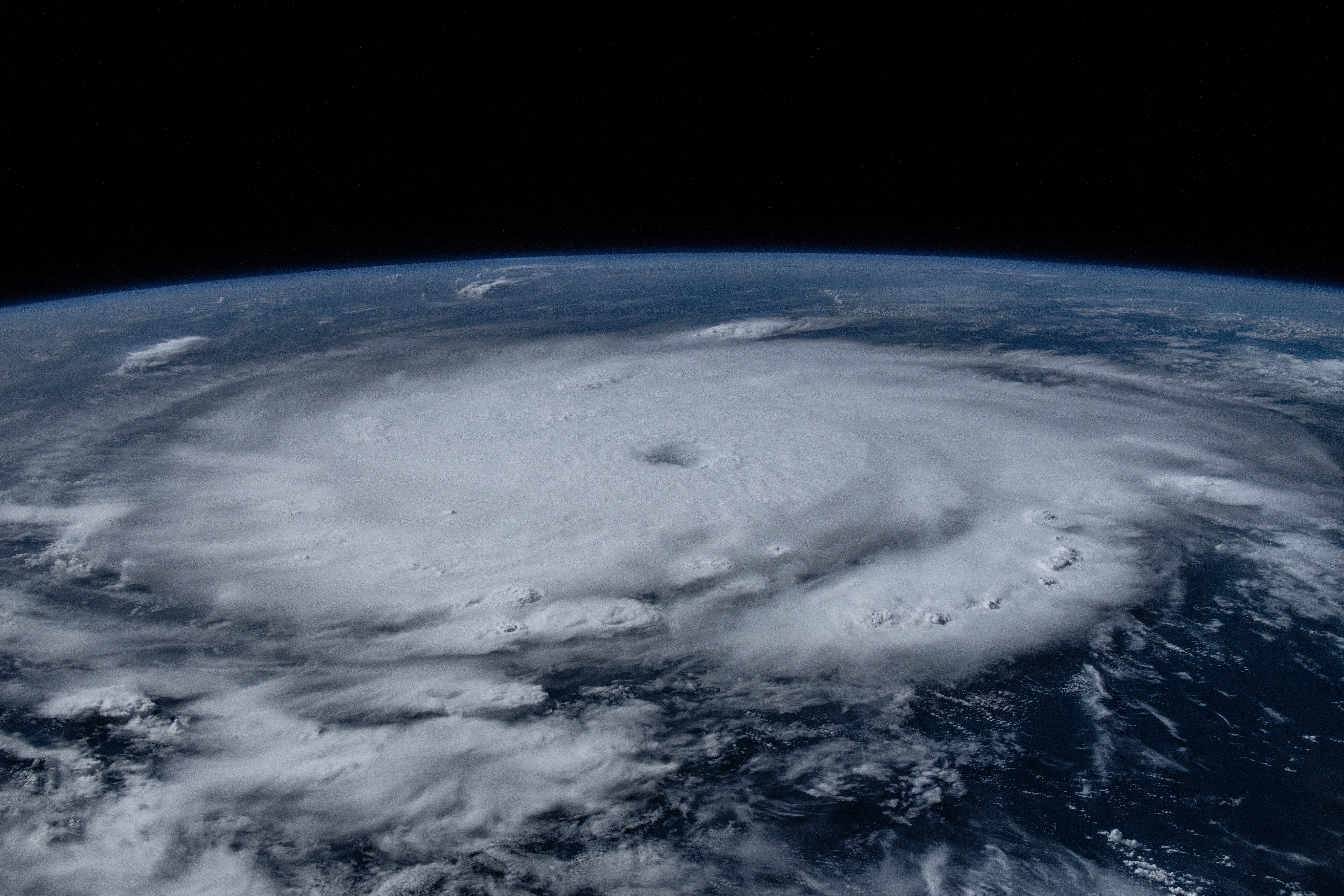A view of Hurricane Beryl from the International Space Station. The hurricane is a big white circle of clouds, with spiral arms visible at far right and bottom middle. The surrounding water is various shades of blue: lighter blue at the top and deeper blue at the bottom of the photo. Earth's curve is visible in the back, up against the darkness of space.