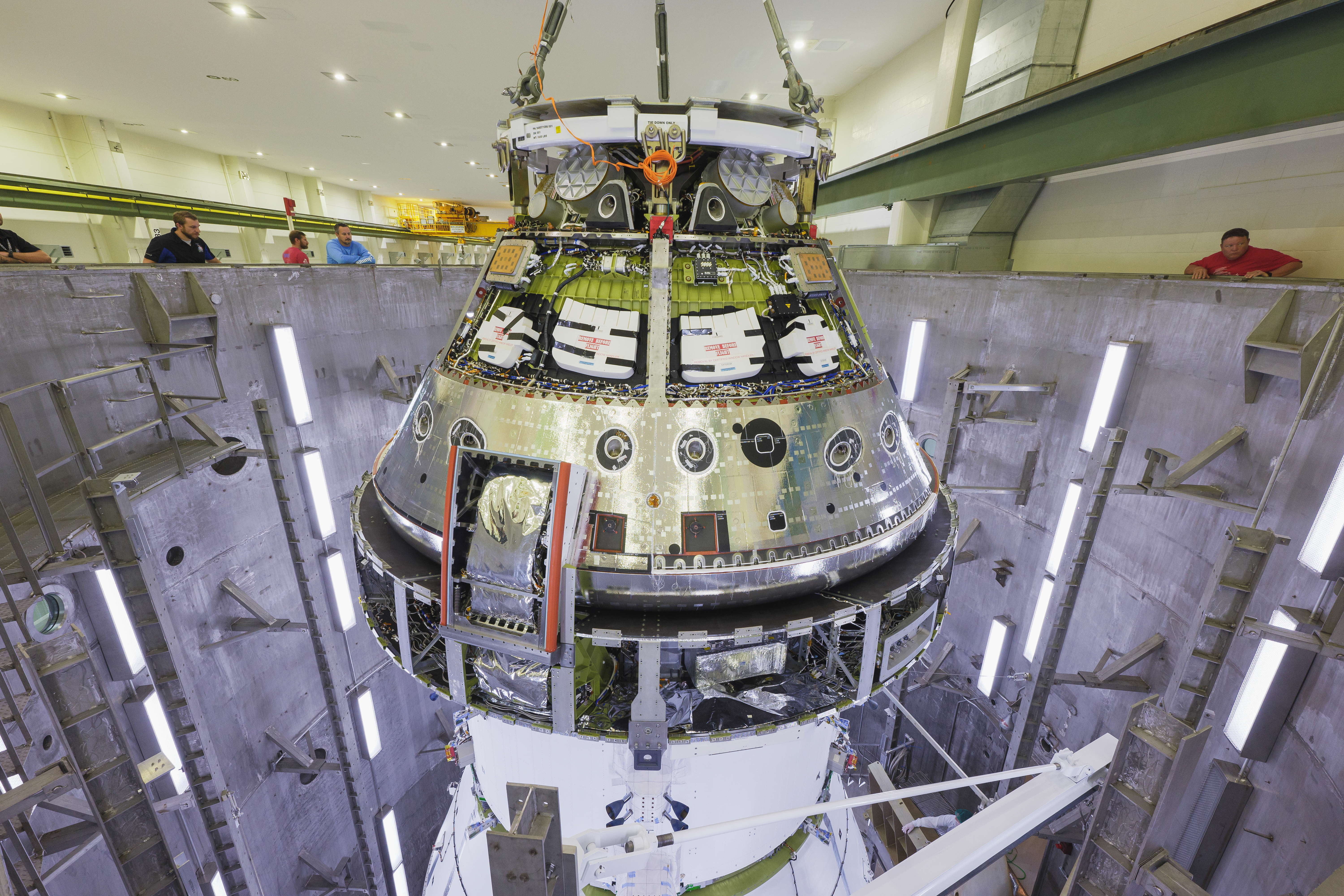 The Artemis II Orion spacecraft is pictured surrounded by the metal walls of the altitude chamber. Orion is a cone shaped spacecraft with metal and wires exposed. Technicians stand around the open top of the altitude chamber.