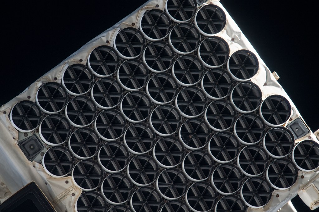 : NICER’s X-ray concentrators are dark circles in eight staggered rows covering this image. Each one is divided into six segments, like a sliced pie, by its sunshade. The concentrators rest in a white frame of the telescope.