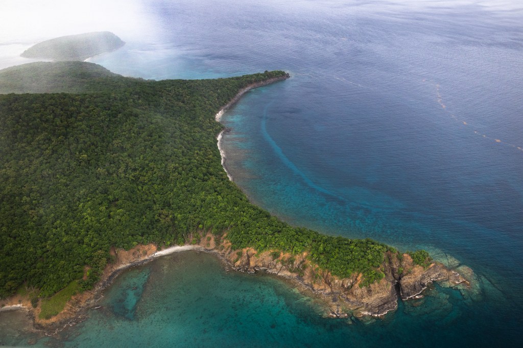 A green forested island sits in bright blue water, that turns teal as it touches land. IN the tip left of the picture, a white haze shows where rain is coming down.