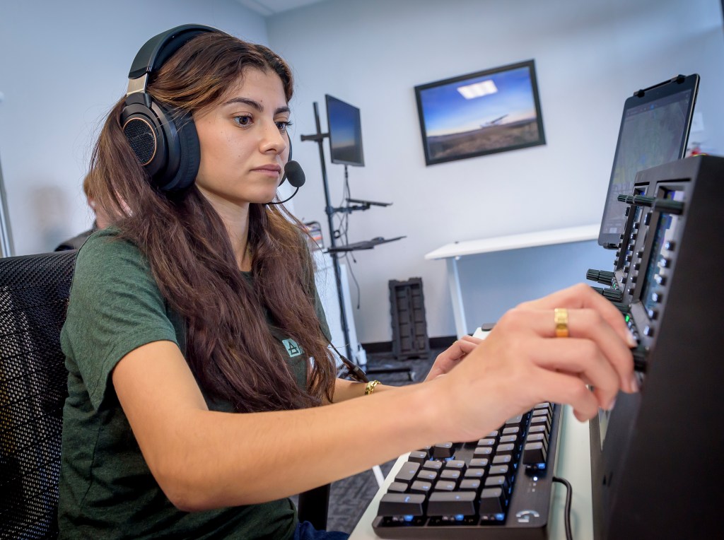 A woman with long brown hair wearing a green t shirt and over the ear headphones is sitting at a desk. She reaches forward with her right hand to adjust a control in front of her.