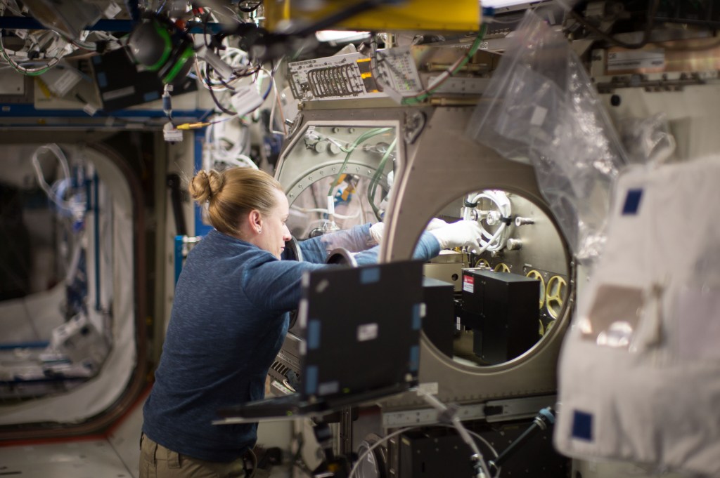 An astronaut works inside the International Space Station, focused on an enclosed laboratory experiment. She wears a blue sweater and gloves and carefully interacts with equipment inside a metallic chamber.