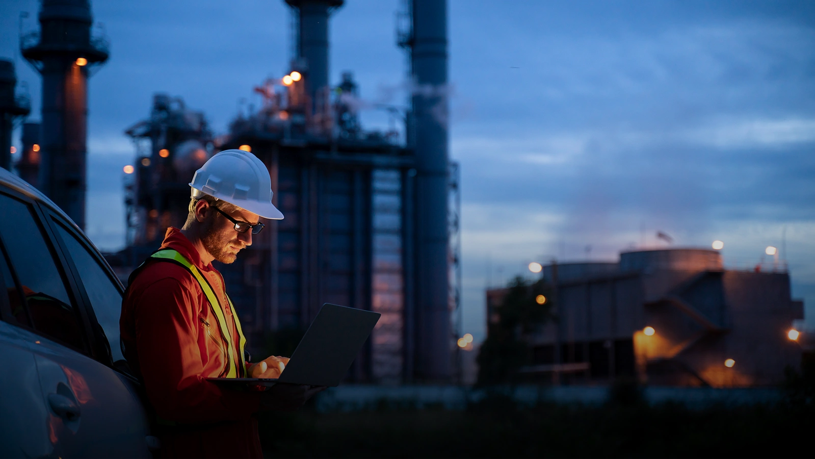 Construction worker leaning against vehicle at dusk, with tall buildings in the background