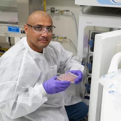 A researcher wearing safety gear kneels at the open door of a lab fridge, displaying a tray of samples. 