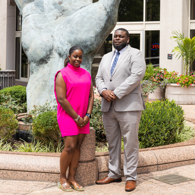 Two people in business formal clothing pose for a photo outside in front of a large statue of a hand.