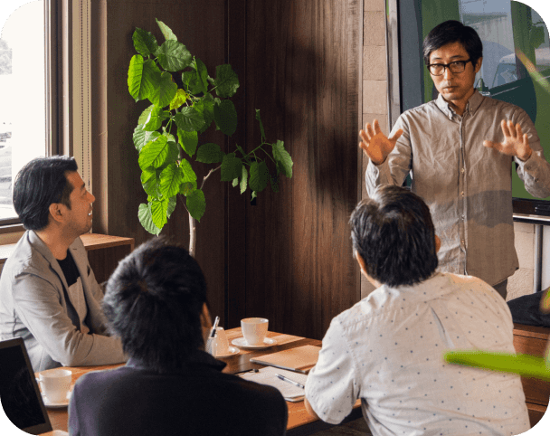 Man standing making hand gestures in meeting while three other individuals sit around conference table