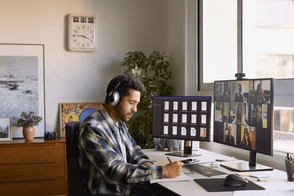 A man sitting in his home office taking notes and smiling softly with a Zoom call on his desktop in front of him