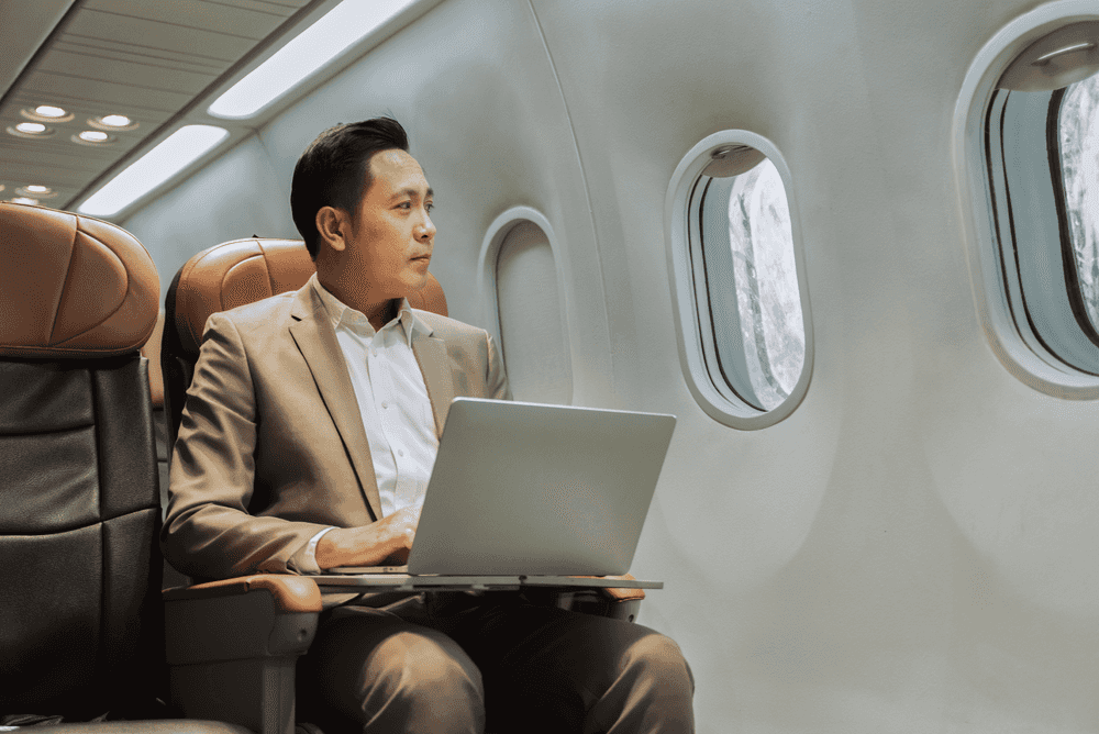 A business man working at his laptop on an airplane is looking out at the sunlight through the passenger window