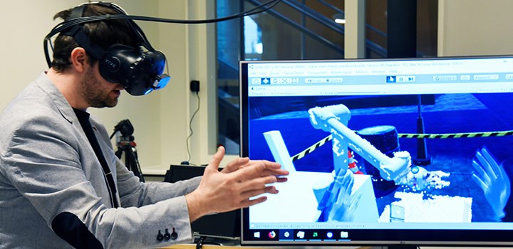 A man wearing VR glasses, with a display in the background showing a robot arm.