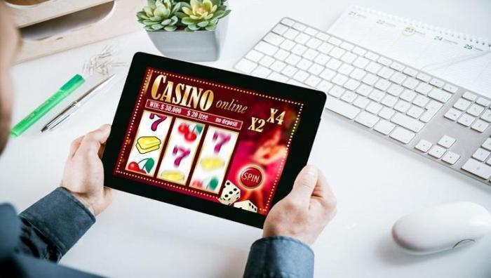 H&auml;nder som h&aring;ller surfplatta med casinospel mot en bakgrund av ett tangentbord, en datormus, en v&auml;xt, pennor med mera