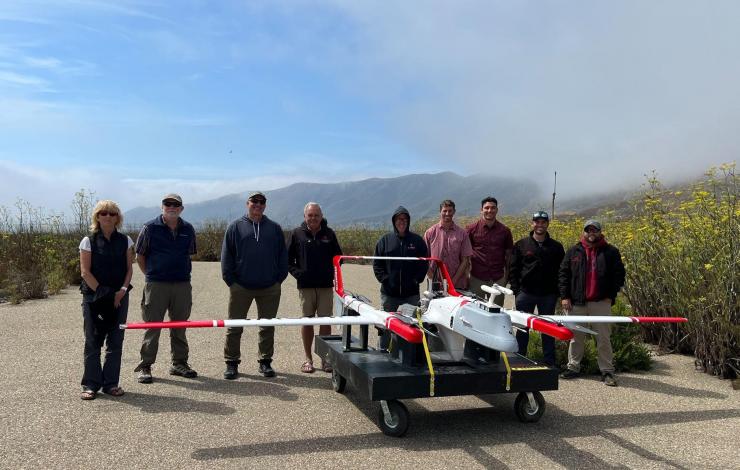 A group of 9 standing next to each other smiling on a partly cloudy sky behind a large white and red striped drone on a runway. 