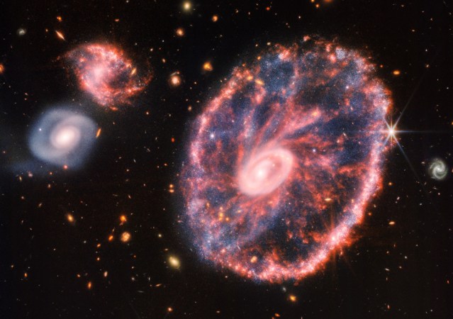 Ring galaxy with two small spiral galaxies to the left in red, blue, and yellow in a James Webb Space Telescope composite image