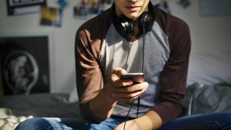 Teen boy with headphones in bedroom listening to music on smartphone