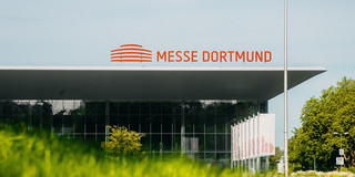 Aufnahme eines großen Gebäudes, auf dem Dach die Aufschrift "Messe Dortmund". Im Vordergrund ist verschwommen eine Wiese zu sehen.