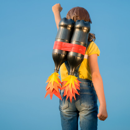 Man sieht eine junge Person mit gelbem T-Shirt, Jeans und Lederkappe von hinten vor einem blauen Himmel. Sie hat eine Attrappe eines Raketenrucksacks auf dem Rücken und streckt die linke Faust in die Höhe.