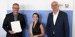 Foto: Eine Frau in schwarzem Kleid und zwei Männern in Anzüge mit Brillen stehen vor einem Banner der Landesregierung Nordrhein-Westfalen. Der linke Mann hält eine Urkunde in der Hand.