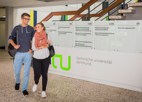 Ein Studentenpärchen läuft an einem TU-Logo im Foyer eines Gebäudes vorbei.