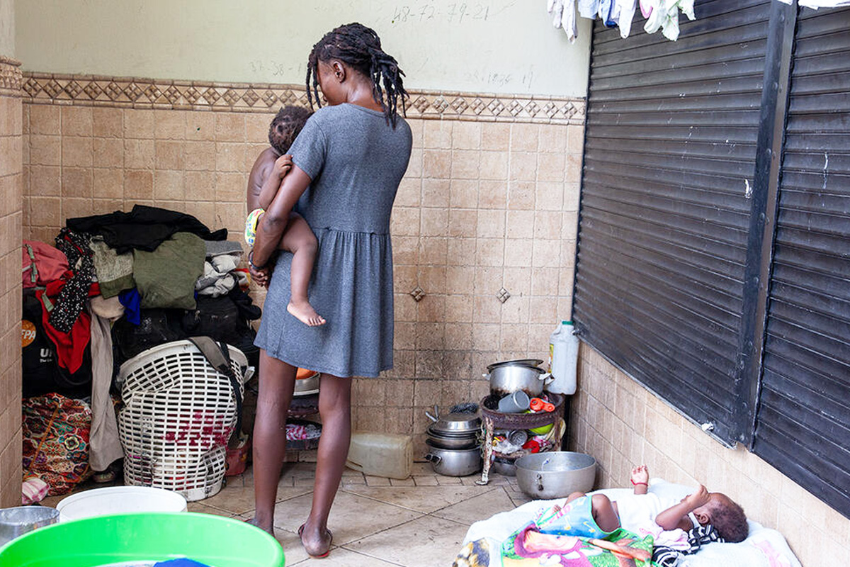 A young woman is seen from the back cradling a toddler while another baby lies at her feet inside a bucket. 