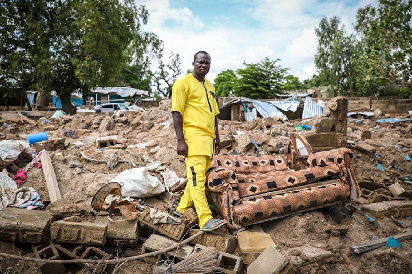 Un homme africain se tient au milieu des ruines d’une maison effondrée, près d’un fauteuil endommagé, avec des arbres en arrière-plan