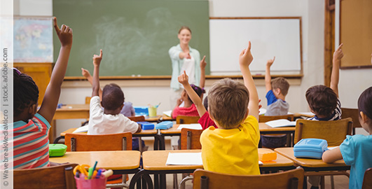 In einem Klassenzimmer sitzen Schülerinnen und Schüler und melden sich, während die Lehrerin vorne eine von ihnen dran nimmt