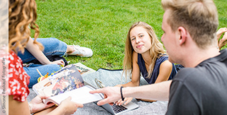 Vier Studierende sitzen auf einer Decke im Schlossgarten, eine von ihnen schaut dem Betrachter entgegen.