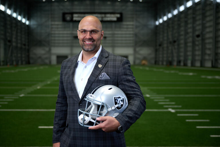 man standing on indoor football field holding Las Vegas Raiders helmet